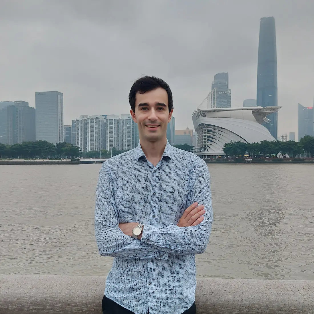 Alex in a patterned shirt standing by the riverside with arms crossed and a confident smile, city skyline in the background.
